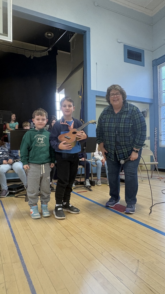 Two boys stand beside an adult woman as one boy holds a small ukulele award during a school assembly.
