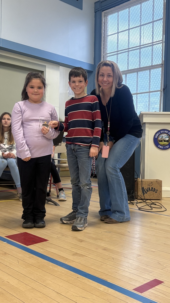 Two children stand next to an adult woman, holding a small award together in a school gym.