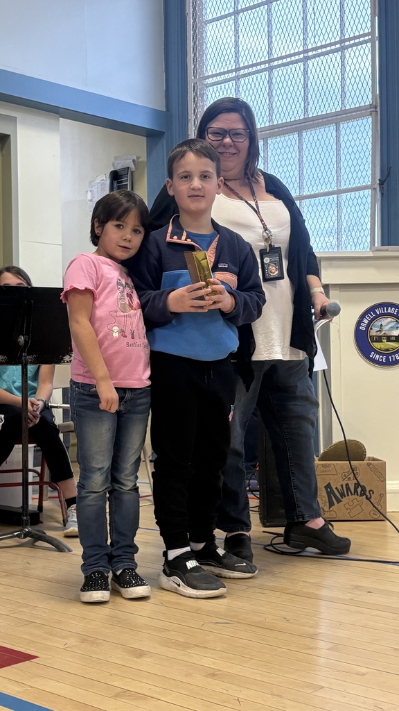 A young boy stands with a young girl and an adult woman, holding a gold-wrapped award in a school gym.