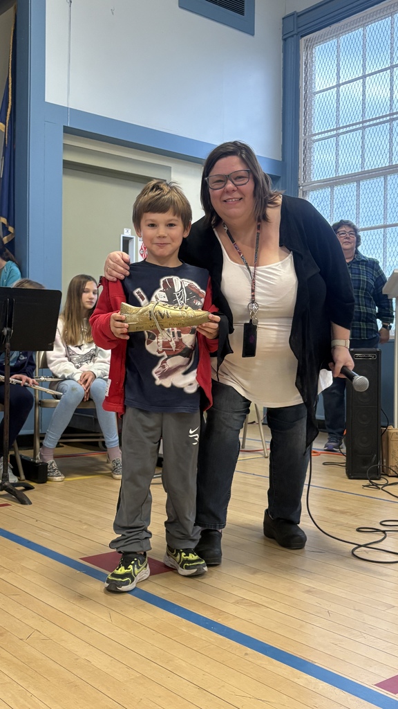 A smiling young boy stands beside an adult woman holding a golden shoe award during a school assembly.