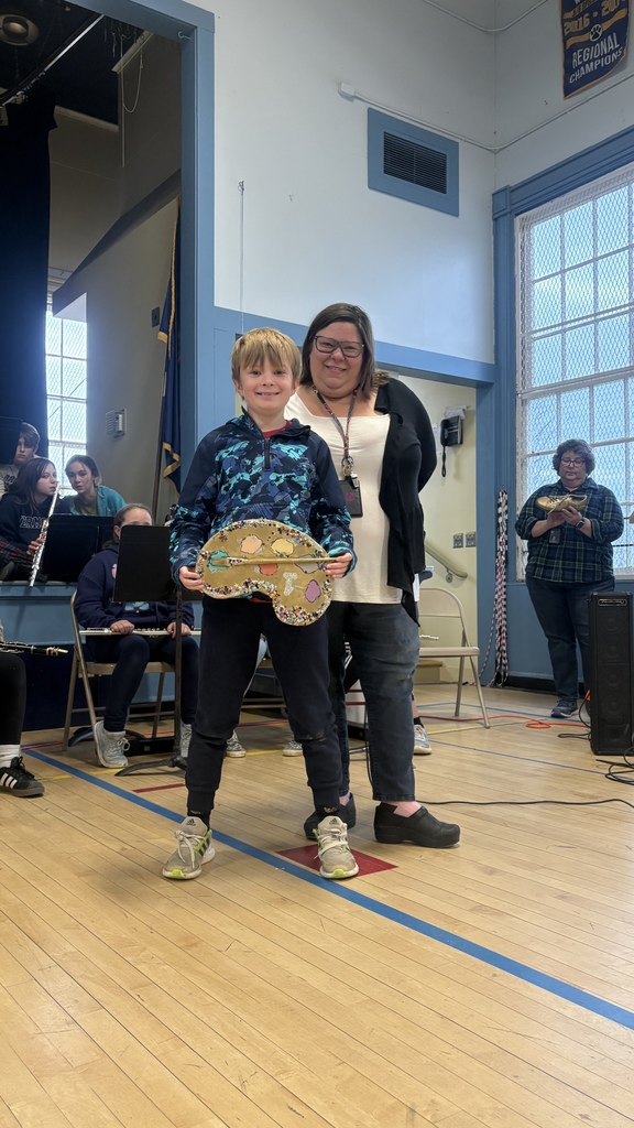 A young boy stands with an adult woman while holding a decorated artist’s palette award during a school assembly.