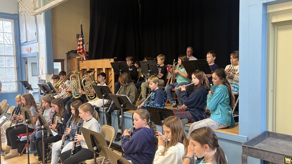 A group of 5th–6th grade band students play brass and woodwind instruments, with Principal Walters seated behind them on the drums.