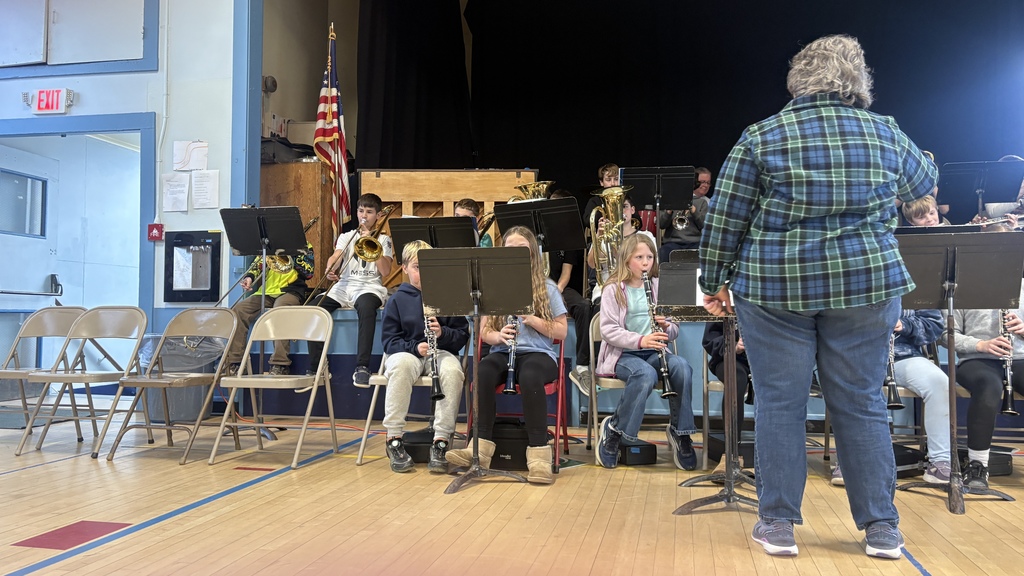 Students in the 5th–6th grade band play instruments onstage while the band director stands in front conducting.
