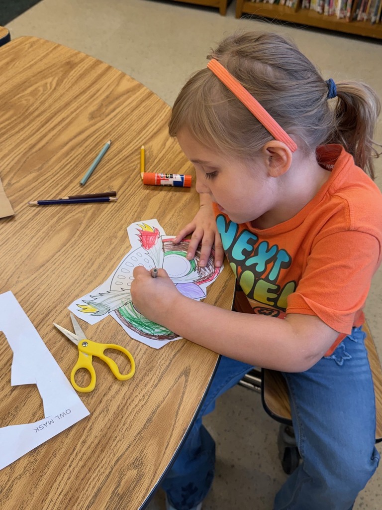 A girl colors her owl mask template with markers at a classroom table.