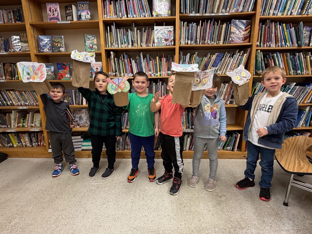 A group of kindergarten students stand in the library holding up their colorful paper-bag owl puppets and smiling.