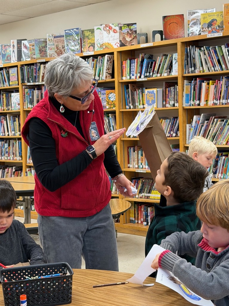 A teacher in a red vest talks with students as they work on coloring and assembling owl puppets.