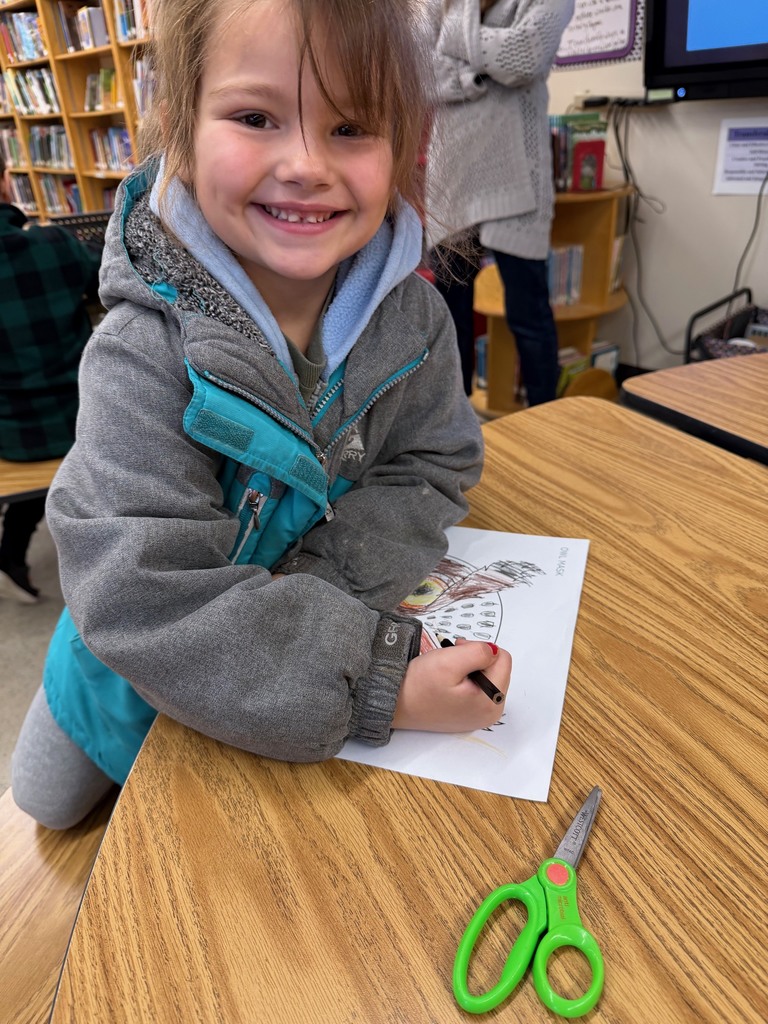 A girl smiles while coloring her owl mask at a classroom table.