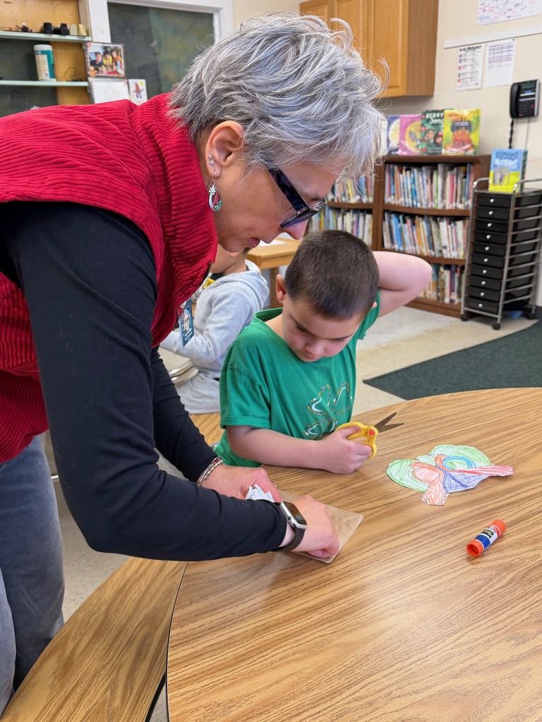 A teacher helps a student glue pieces onto his owl puppet at a classroom table.