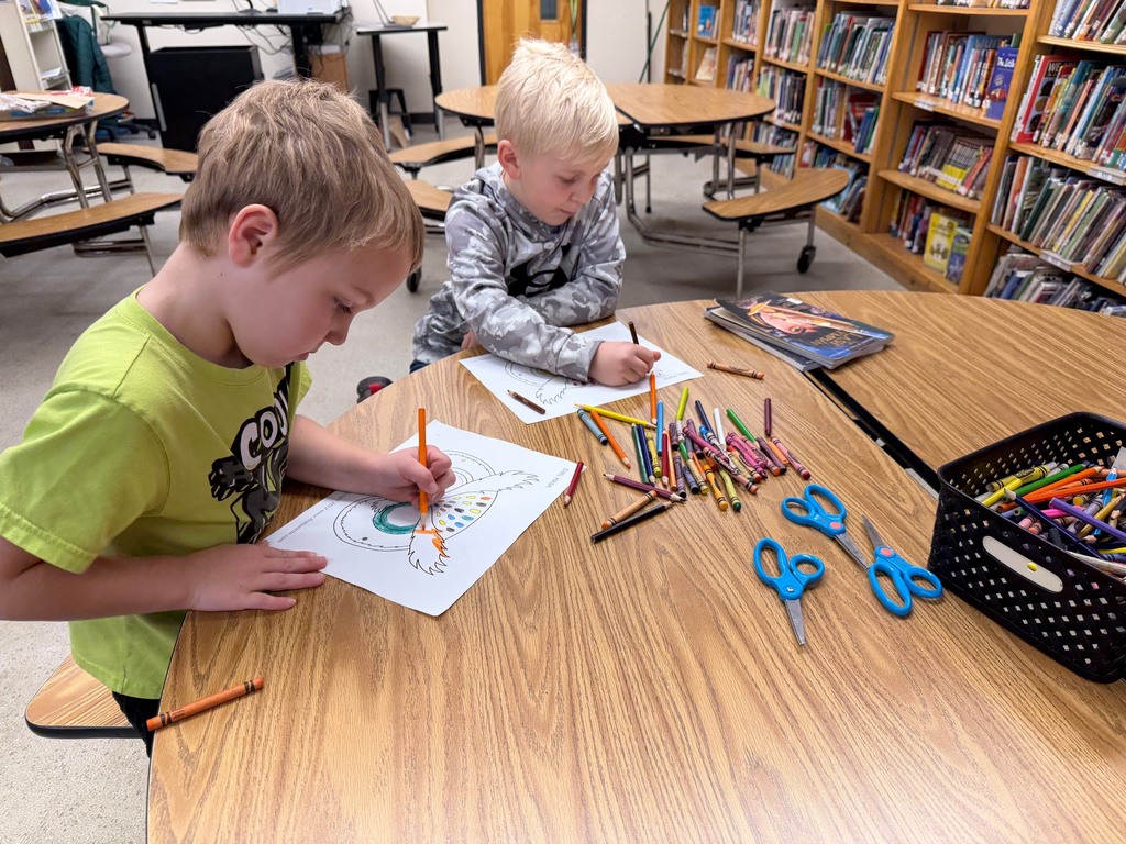 Two students color owl masks with crayons at a round classroom table.