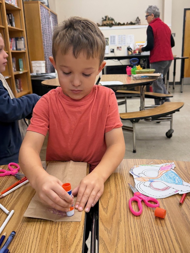 A young boy sits at a table gluing pieces onto his brown paper owl puppet.