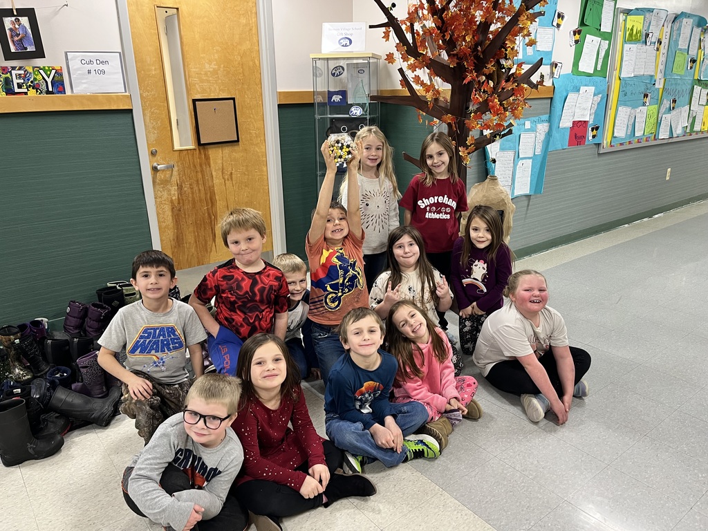 A group of about fifteen young children sit and kneel together in a school hallway, smiling for the camera. One child in the center stands and holds up a small trophy or object. Behind them is a door labeled “Cub Den #109,” a fall-themed decorative tree with orange leaves, and a bulletin board displaying student work. Several pairs of boots are lined up along the wall to the left.