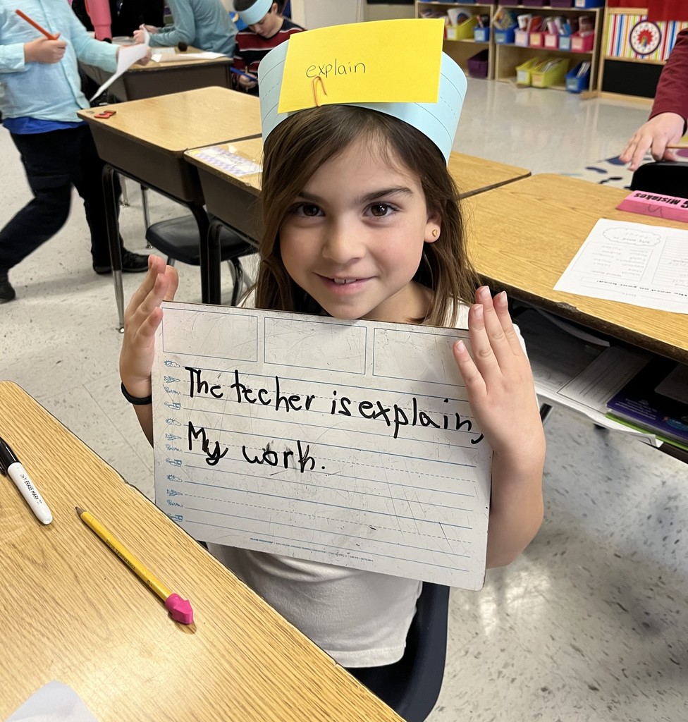  A young elementary school girl with long dark hair, wearing a blue and yellow paper crown with the word "explain" written on it. She is smiling and holding a dry-erase board that says, "The teacher is explaining my work.