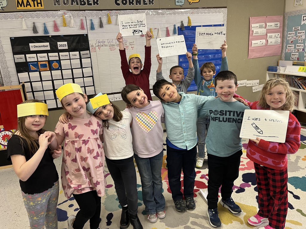  (Large group of children): A large group of elementary school children smiling in a classroom. The four girls from the previous photo are on the left, and three boys are in the back holding up dry-erase boards. The boy on the far right is holding a board with a drawing of a crayon and the text "I was a crayon."
