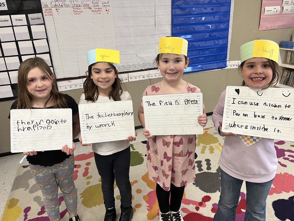 Group of 4 girls): Four elementary school girls smiling and holding dry-erase boards with sentences. They are wearing colored paper crowns with words written on them: 'explain,' 'field,' and 'label.' The sentences read, from left to right: "there is going to be a wrap [or 'wrench']", "The tech is explaining my wrench", "The field is green.", and "I can use a label to put on boxes to know what is inside it." 
