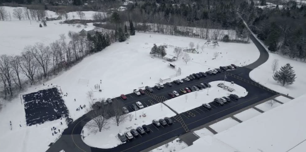 Drone view of a school campus covered in fresh snow, with students playing on the playground and fields. Cars are parked in neat rows along the driveway, and trees surround the snowy grounds.