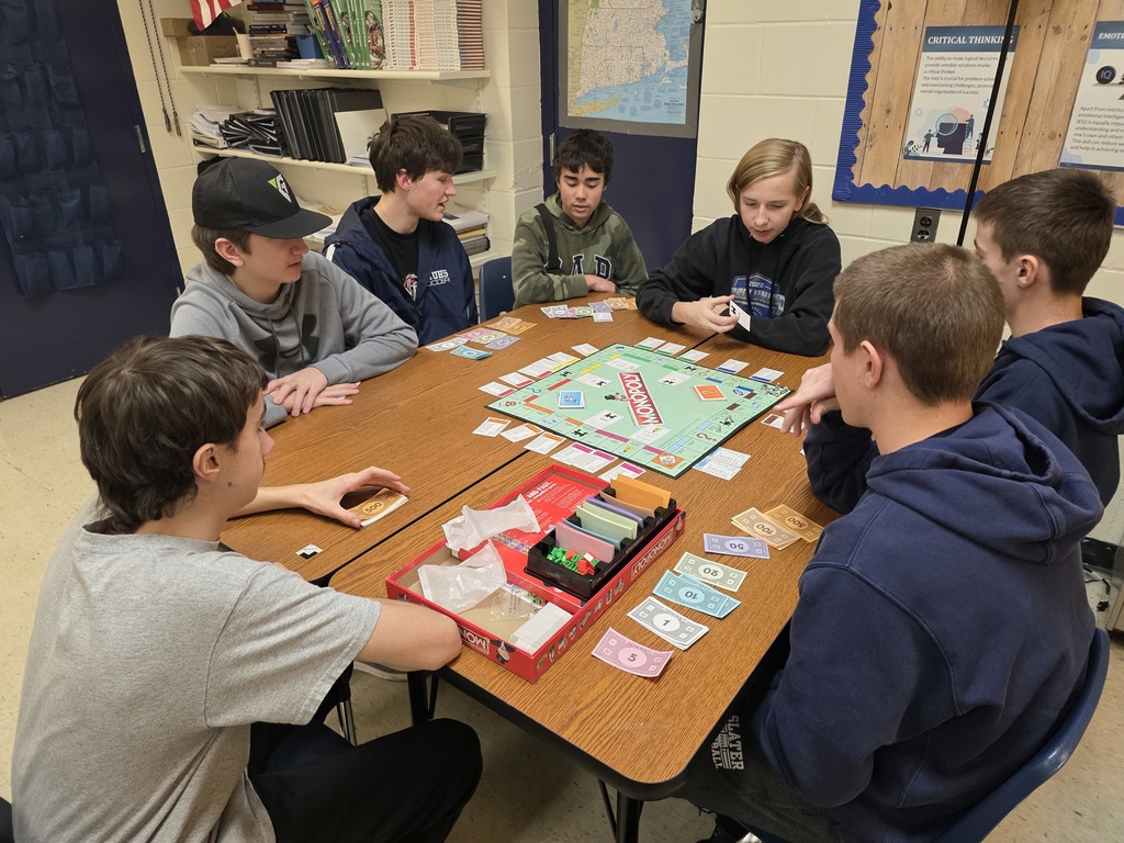 A group of students seated at a table in a classroom playing Monopoly together, with game pieces and money spread out.