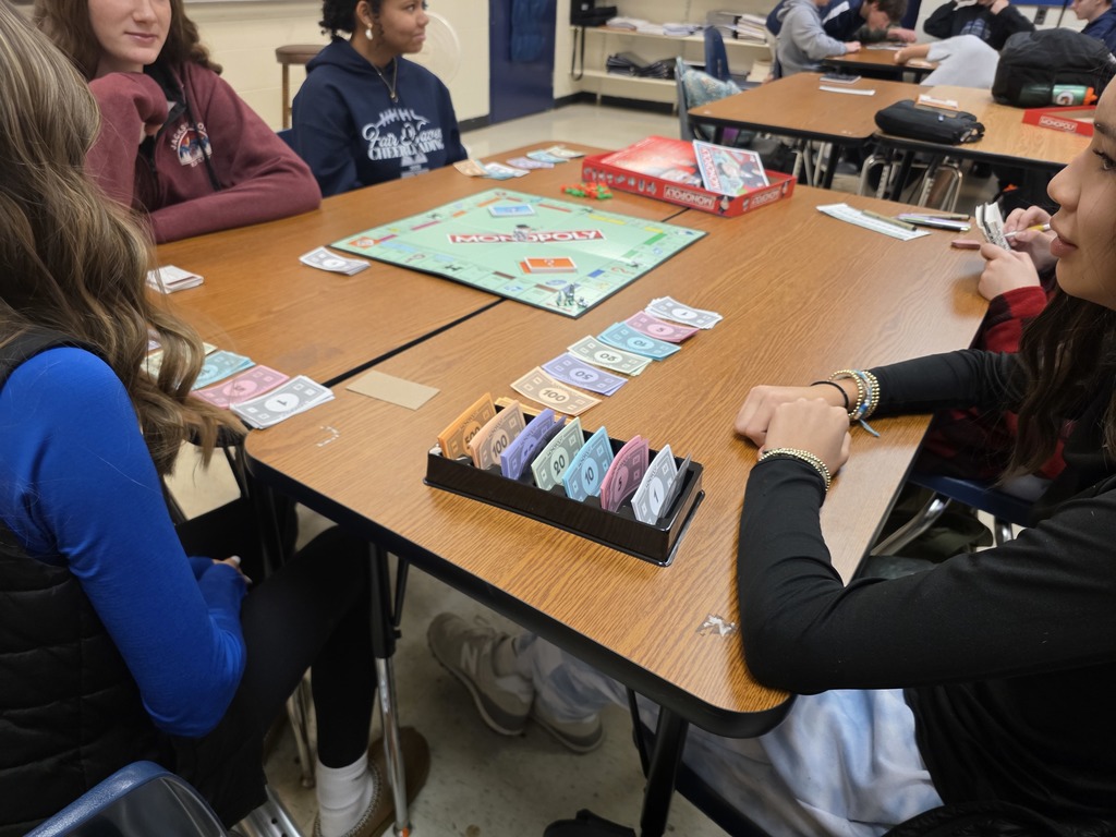 Students sitting around a classroom table playing Monopoly, with game money and cards organized in front of them.