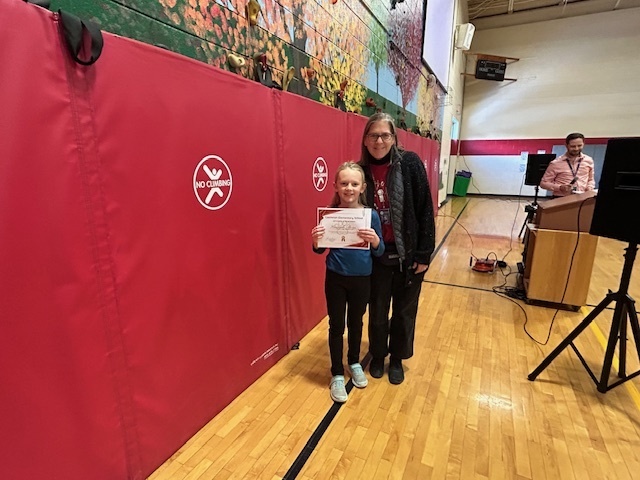 A student holds a certificate while standing next to an adult in the school gym.