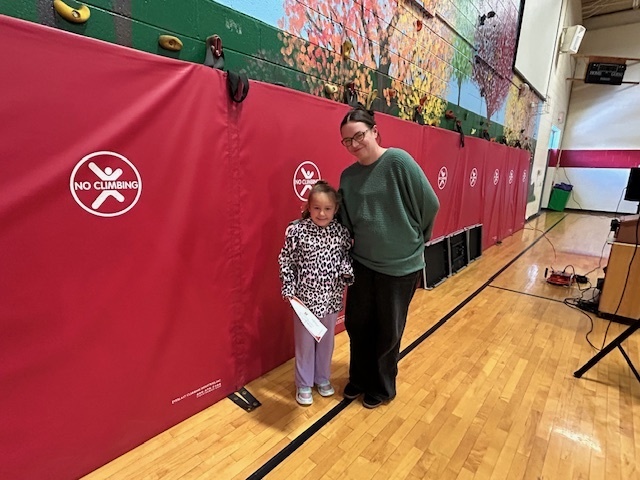 A student holding a certificate stands beside an adult in a gym with red mats.