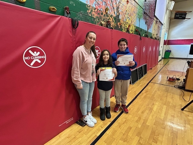An adult stands with two students holding certificates in a gym with red mats.