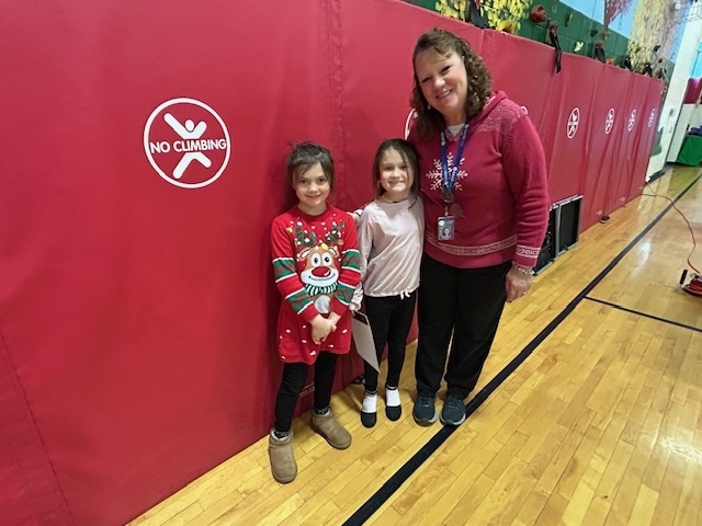 Two students stand with an adult in front of red gym mats; one student holds a certificate.