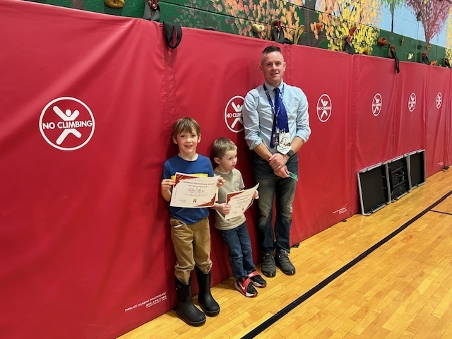 Two young students hold certificates while standing beside an adult in a gym with red wall mats.