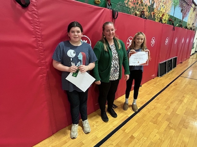 Two students stand with an adult in front of red gym mats; one holds a certificate.
