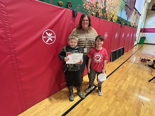 Two boys hold certificates while standing with an adult in front of red gym mats.