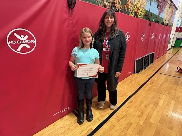 A student holding a certificate stands with an adult in front of red mats in the gym.