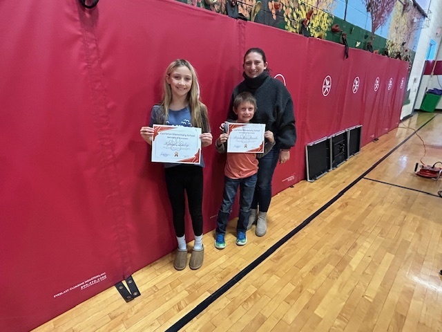Two students hold certificates while standing with an adult in front of red gym wall pads.
