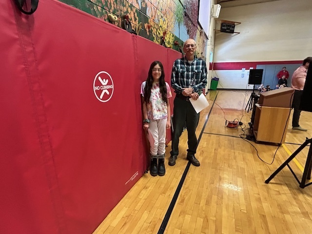 Student stands with an adult near red gym wall pads, holding a certificate.