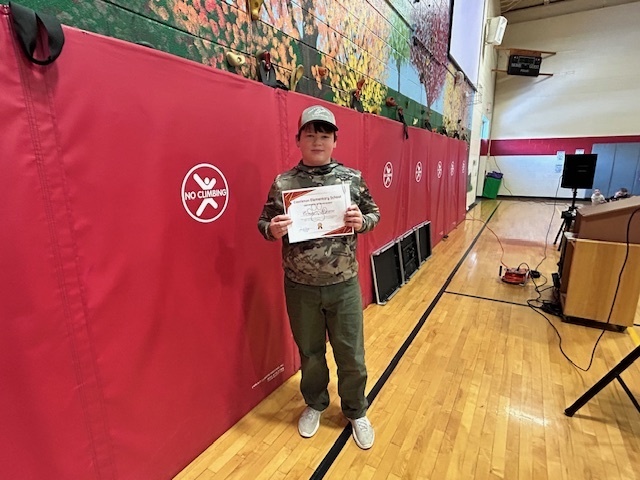 Student stands with an adult in front of red gym wall pads, holding a certificate.