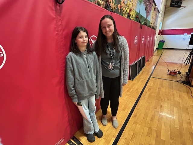 Student stands with an adult in front of red gym wall pads during an awards presentation.