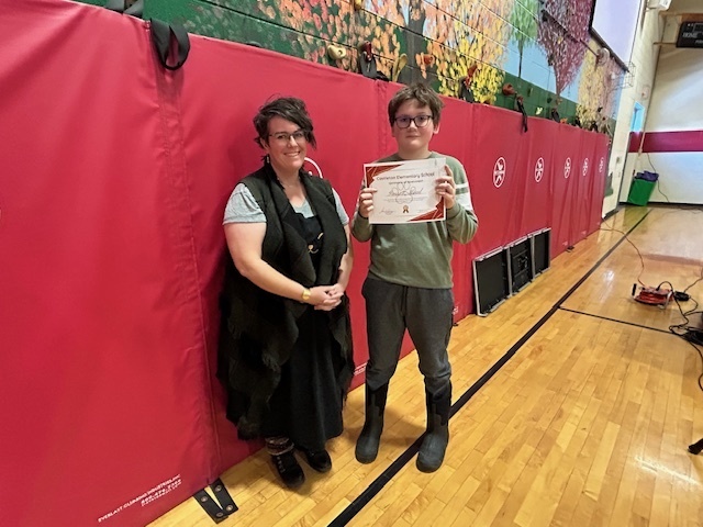 Student stands beside an adult, holding a certificate, in front of red gym wall pads.