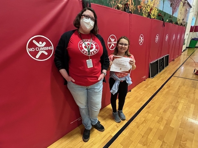 Student stands with an adult holding a certificate in front of red gym wall pads.