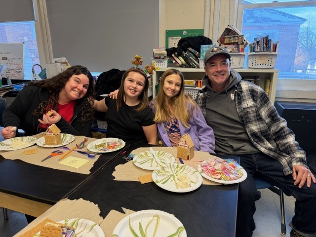 A family of four smiling at a table with assembled gingerbread houses and candy decorations.