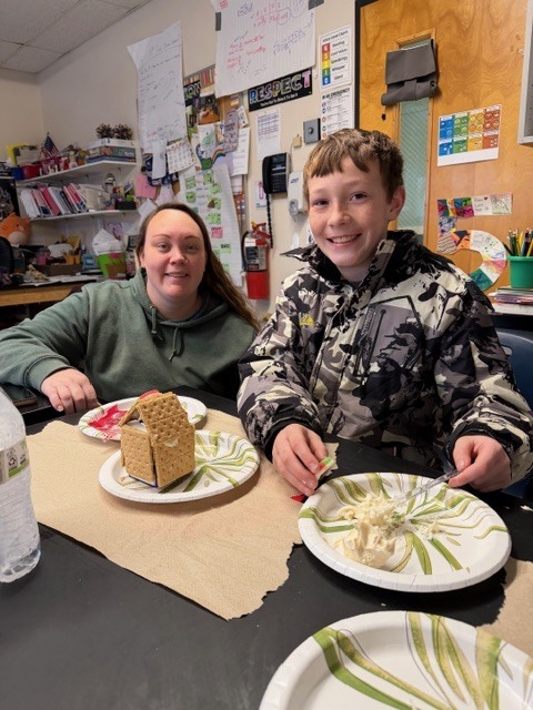 A student and an adult smiling at a table with a partially built gingerbread house on a paper plate.