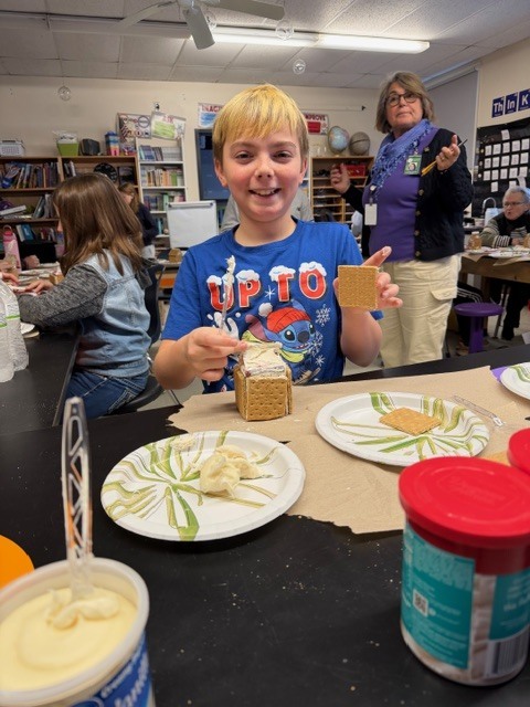 A smiling student holding graham crackers while building a gingerbread house, with frosting and supplies on the table.