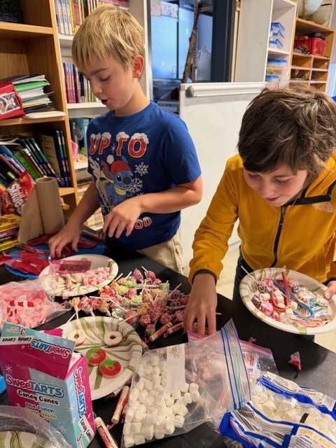 Two boys selecting candy decorations from a large assortment spread across a table.