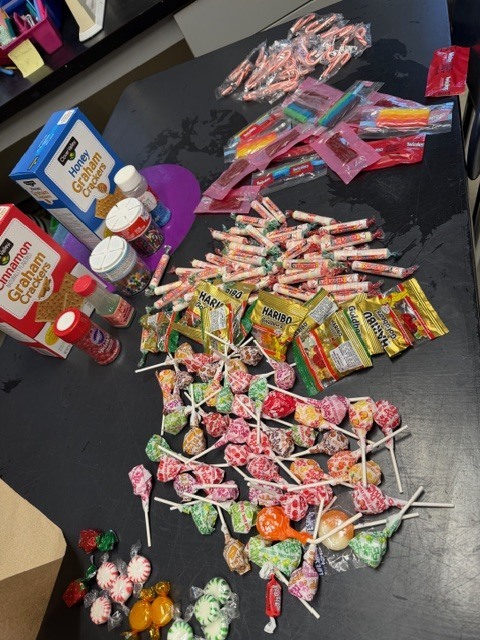 A table covered with assorted candy, including lollipops, Smarties, gummies, and sprinkles, for decorating gingerbread houses.