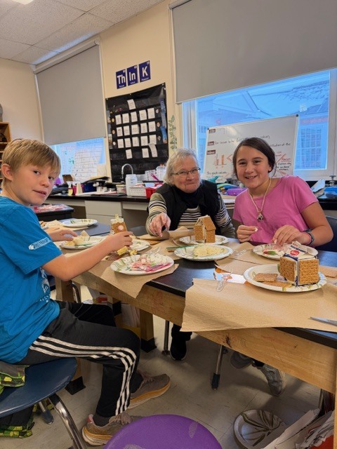 Three people—an adult and two students—working on gingerbread houses at a classroom table.