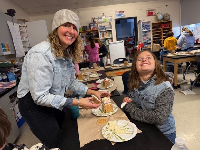 An adult and a student smiling while assembling a gingerbread house at a classroom table.
