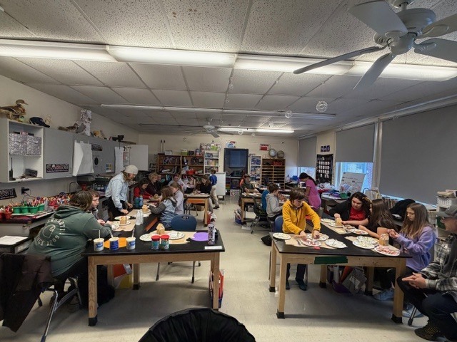 A classroom filled with 5th graders and families sitting at tables, working on gingerbread houses together.