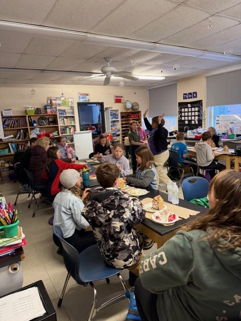Students and families gathered around tables in a classroom, decorating gingerbread houses.