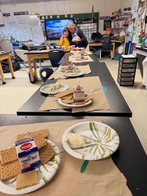A long classroom table set with plates, graham crackers, frosting, and small milk cartons prepared for gingerbread house building.