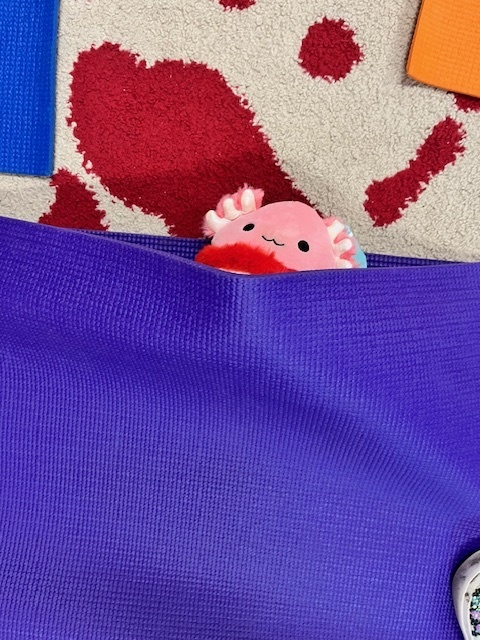 A pink and red plush toy (looks like an axolotl) is peeking out from under a folded purple yoga mat on the floor. A red heart-shaped design is visible on a rug next to the mat.