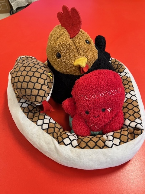 A high-angle photo shows three plush toys sitting together in a small, oval-shaped white bed with a brown, snakeskin-like patterned lining. The largest toy in the back is a brown chicken with a yellow beak, black eyes, and a red comb and wattle. To its left is a plush snake with a brown and black diamond pattern, coiled slightly around the chicken. In the foreground is a bright red, round plush toy with a cheerful embroidered face and a white patterned bandana on its head. The three toys rest on a vibrant red table surface.