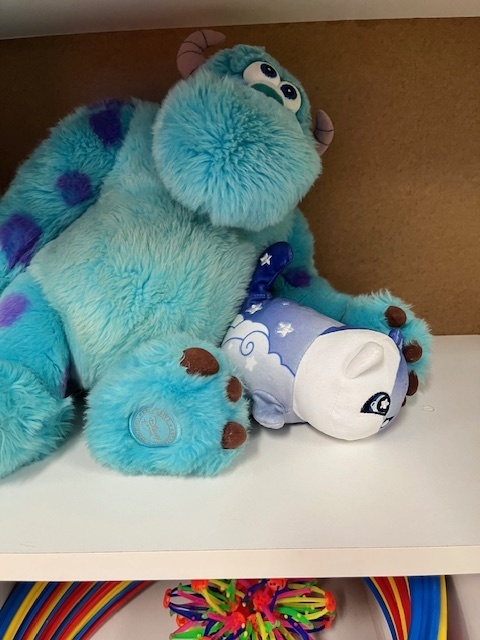. A large, light blue, furry Sulley from Monsters, Inc. is sitting next to a smaller, white and blue sleeping plush toy with a star pattern.