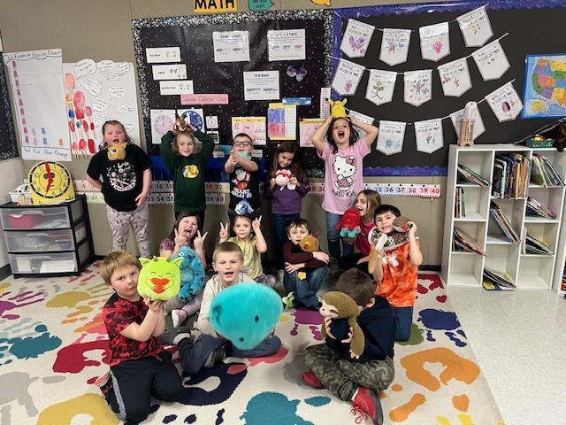 A group of approximately 11 elementary school children, smiling and posing in a classroom while holding various plush toys, including a large blue one, a yellow Pikachu, a green dragon, a sloth, and others. Math and reading decorations are visible on the wall behind them, and they are standing on a colorful splatter-paint rug.