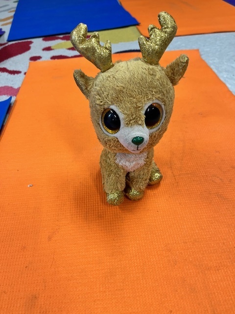 A close-up, centered shot of a small, brown plush reindeer toy with large, sparkly gold eyes and glittery gold antlers, sitting on an orange mat.